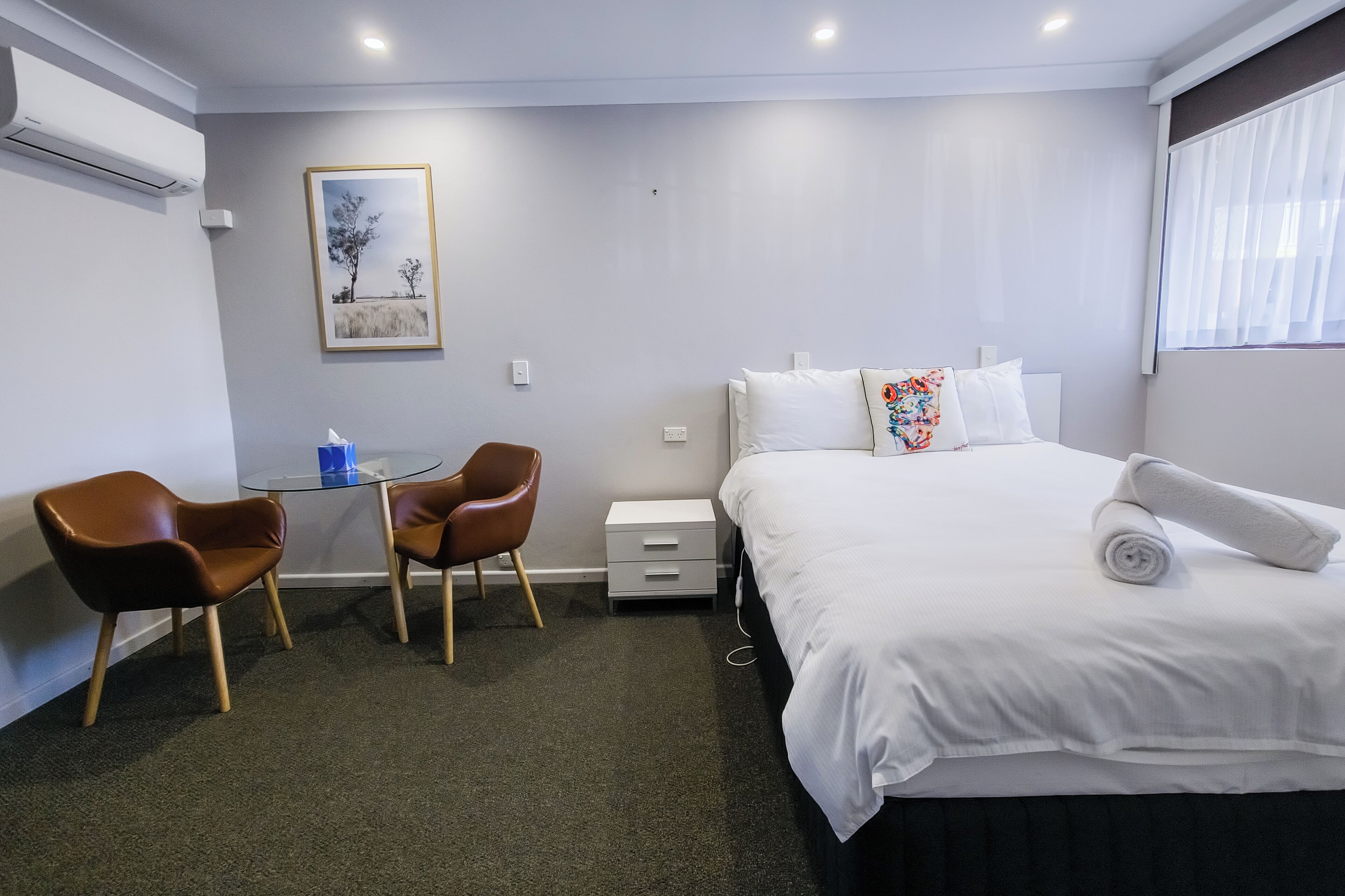 Una habitación de hotel con cama, dos sillas, una mesita, una mesita de noche y un cuadro en la pared.