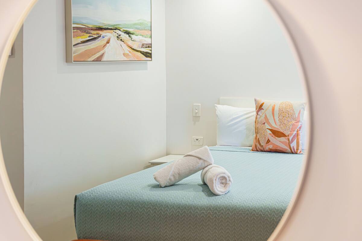 A neatly made bed with a rolled towel, a pillow with a leaf pattern, and a painting on the wall.