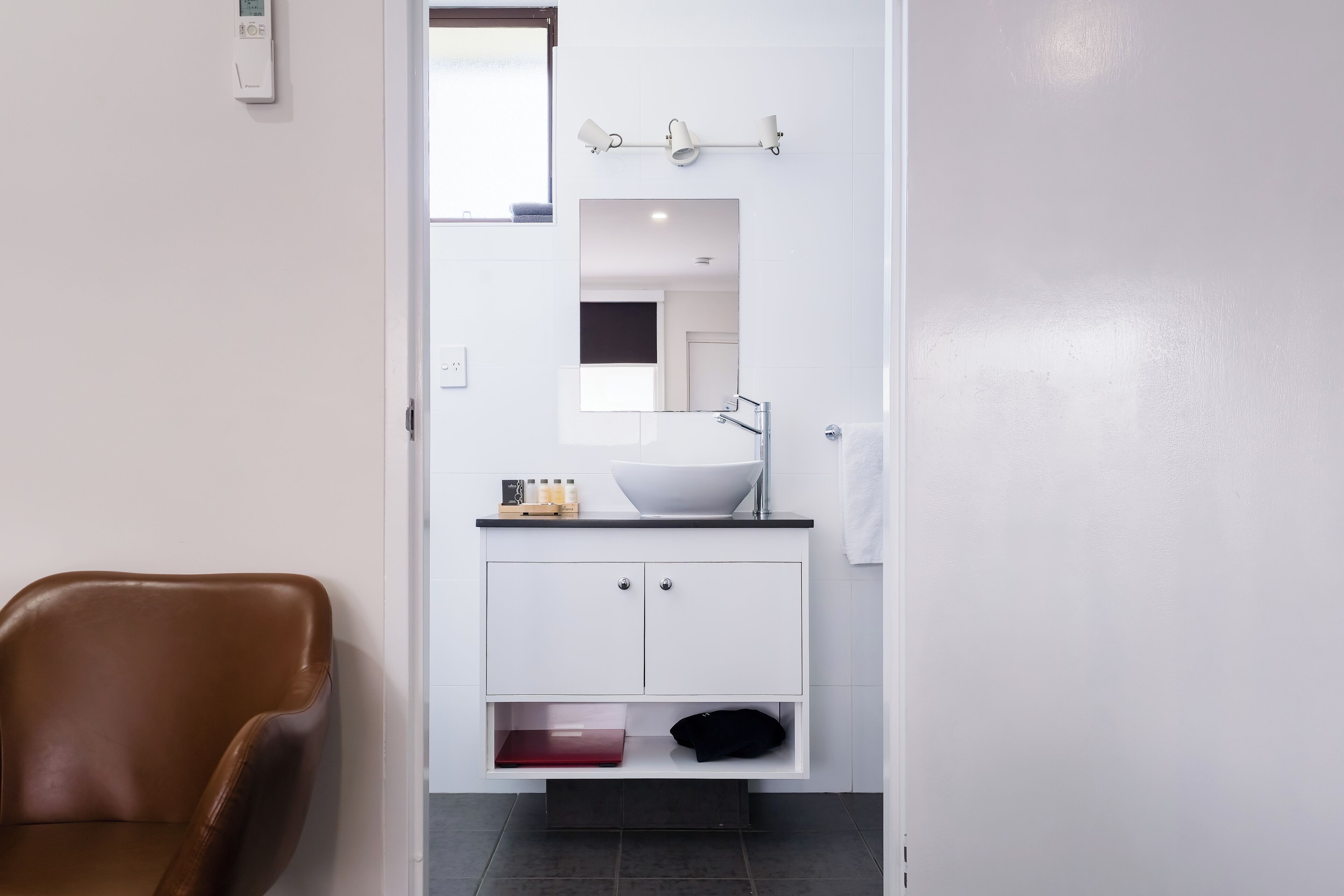 A modern bathroom with a white vanity, a large mirror, and a brown leather chair.