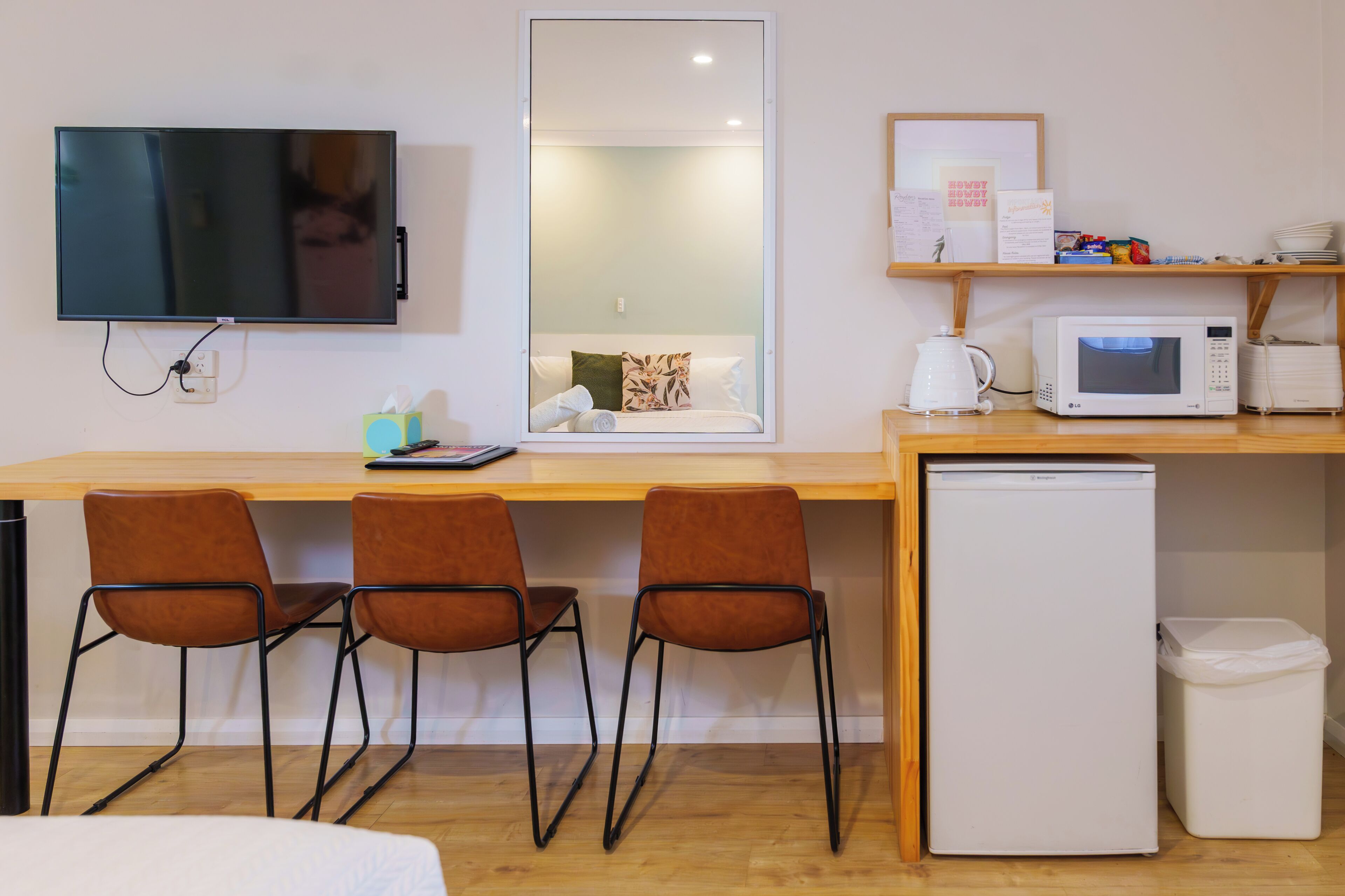 A compact hotel room with a wooden desk, three brown chairs, a television, a microwave, a small refrigerator, and a trash bin.