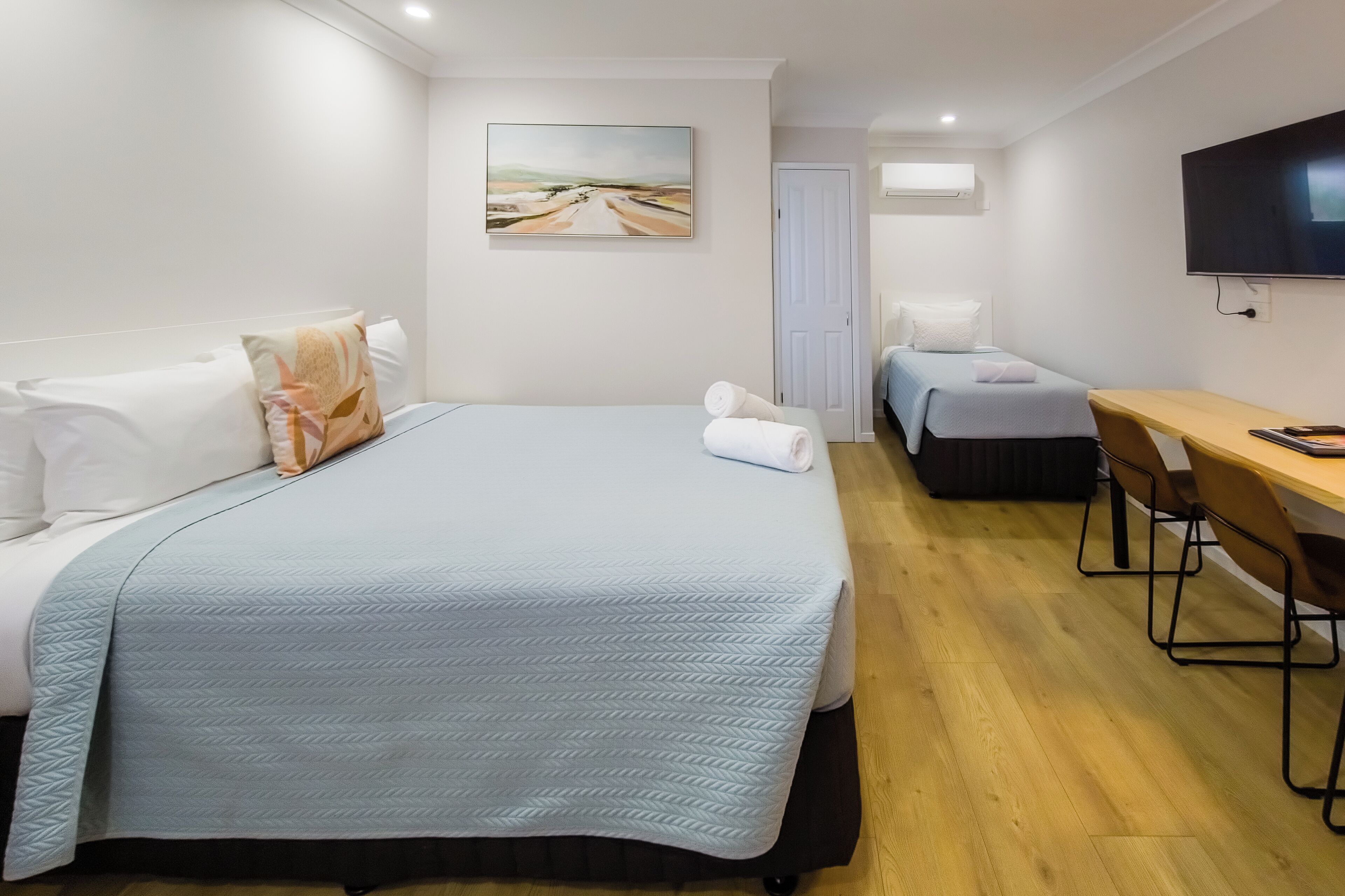 A hotel room with a large bed, a smaller bed, a desk, a chair, a TV, and a painting on the wall.