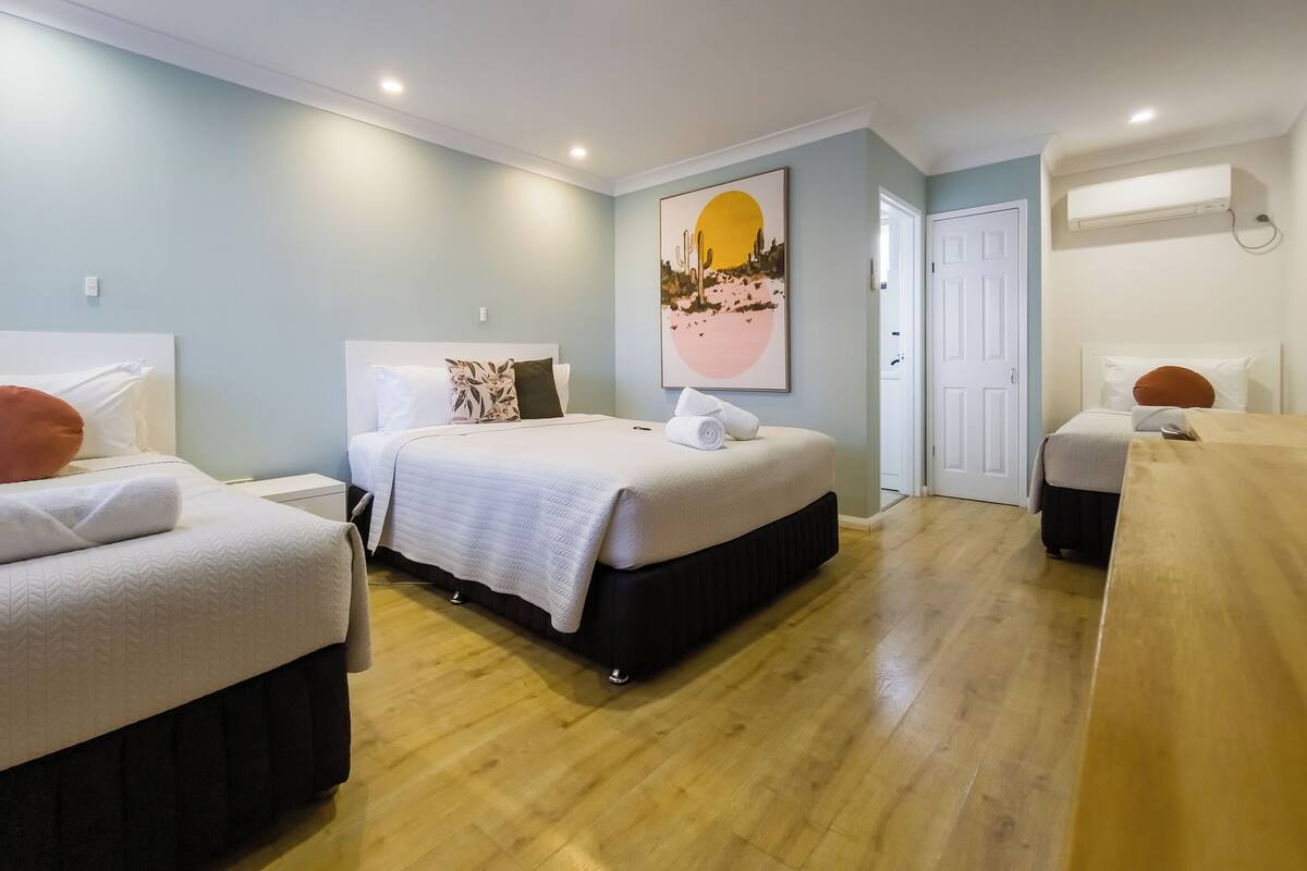 A hotel room with two beds, a wooden floor, a large painting, and an air conditioning unit.