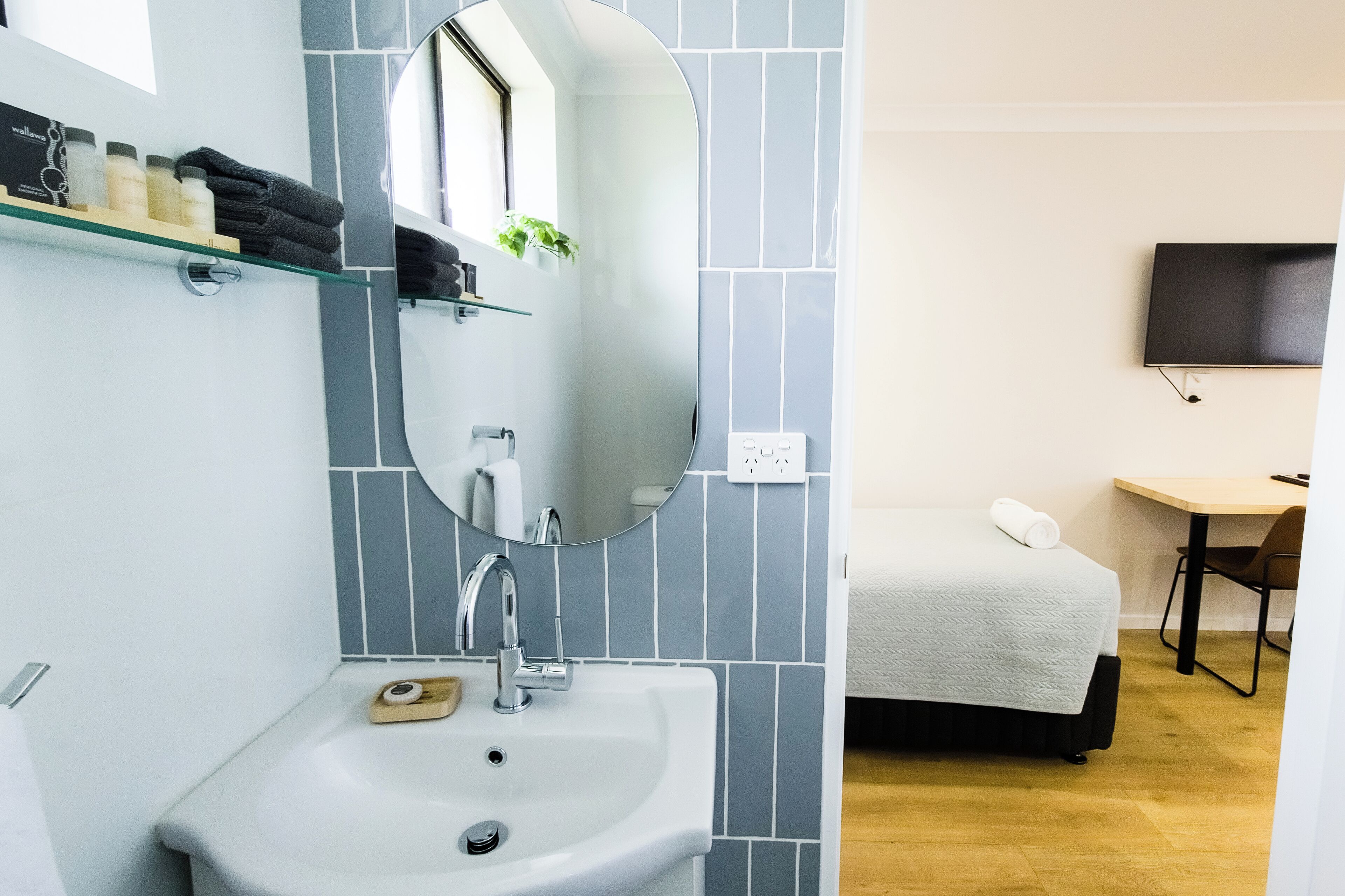 A modern bathroom with a sink, mirror, and toiletries shelf. A bed with a white blanket is visible in the background.