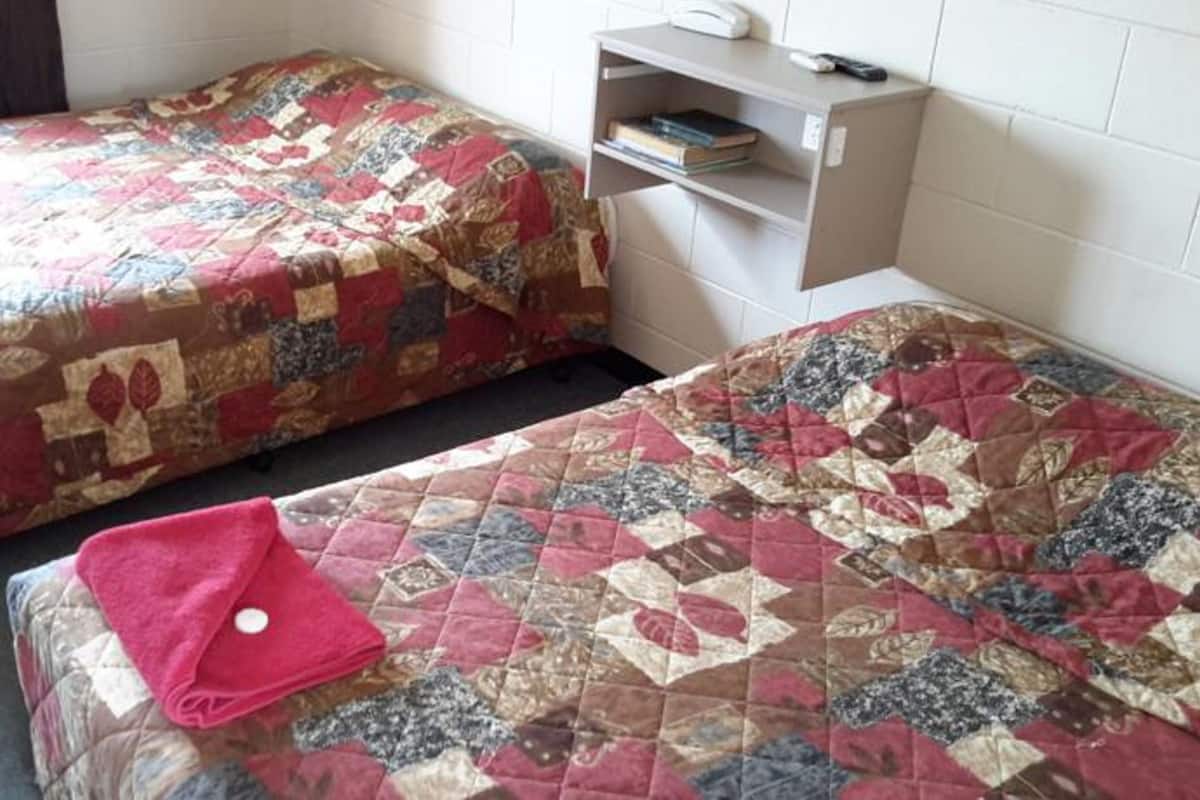 A room with two beds, each with a quilted bedspread. There is a shelf with books and a remote control.