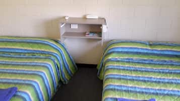 Una habitación con dos camas individuales, cada una con ropa de cama a rayas. Hay un estante pequeño con un teléfono y algunos objetos, además de un enchufe en la pared.