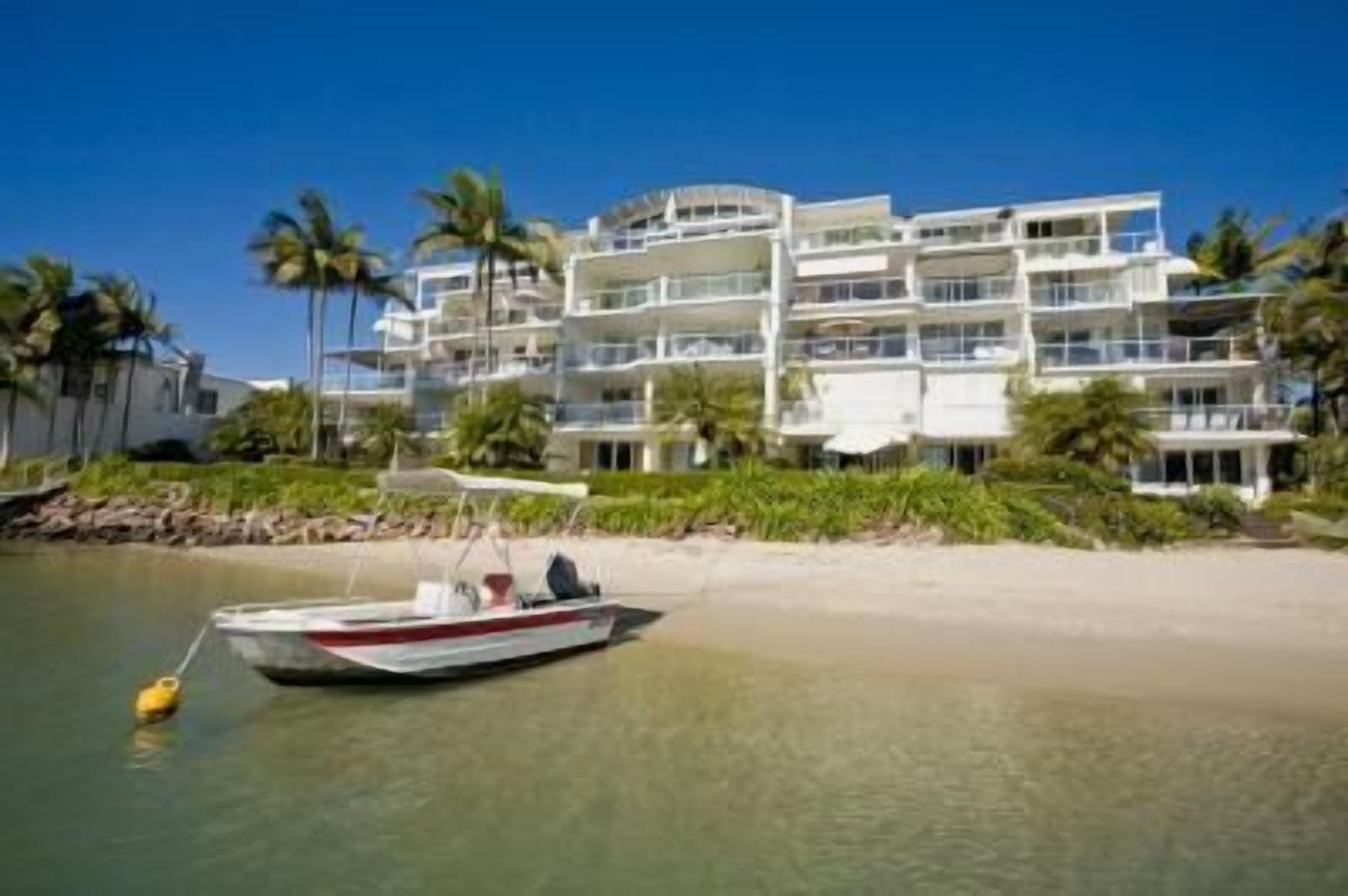 A beachfront hotel with multiple balconies, a boat docked on the shore, and palm trees in the background.