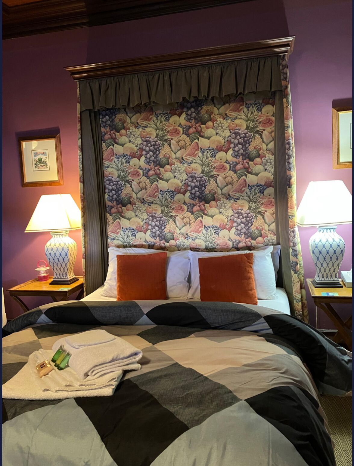 Habitación de hotel con una cama grande, cabecera floral, dos lámparas de noche y un cuadro enmarcado en la pared.