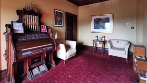 A room with a grand piano, two white armchairs, a red patterned carpet, and framed artwork on the walls. - The Manse (Daylesford)