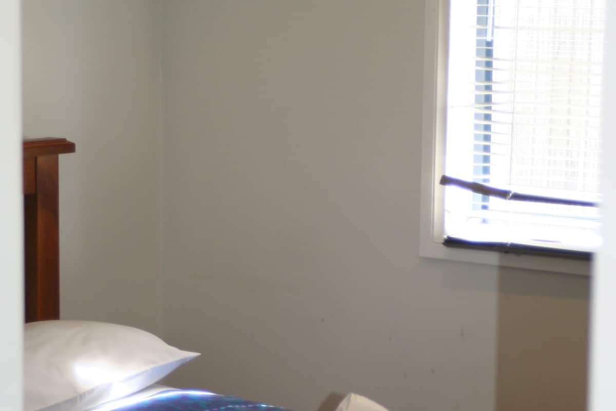 Une petite chambre avec un lit simple, une tête de lit en bois, une serviette enroulée sur le lit et une fenêtre avec des stores.