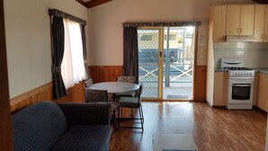 A compact kitchen with wooden cabinets, a gas stove, and a dining area with a table and chairs. - Koonwarra Holiday Park (Lakes Entrance)