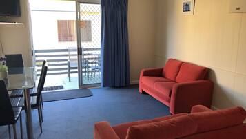 A room with a glass table, two chairs, a red sofa, a TV, and a sliding glass door leading to an outdoor area with a patio and a fence.