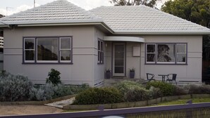 A two-story house with a grey exterior and a tiled roof. There are two windows on the front, a door, and a small porch area with a table and chairs. - Koonwarra Holiday Park (Lakes Entrance)