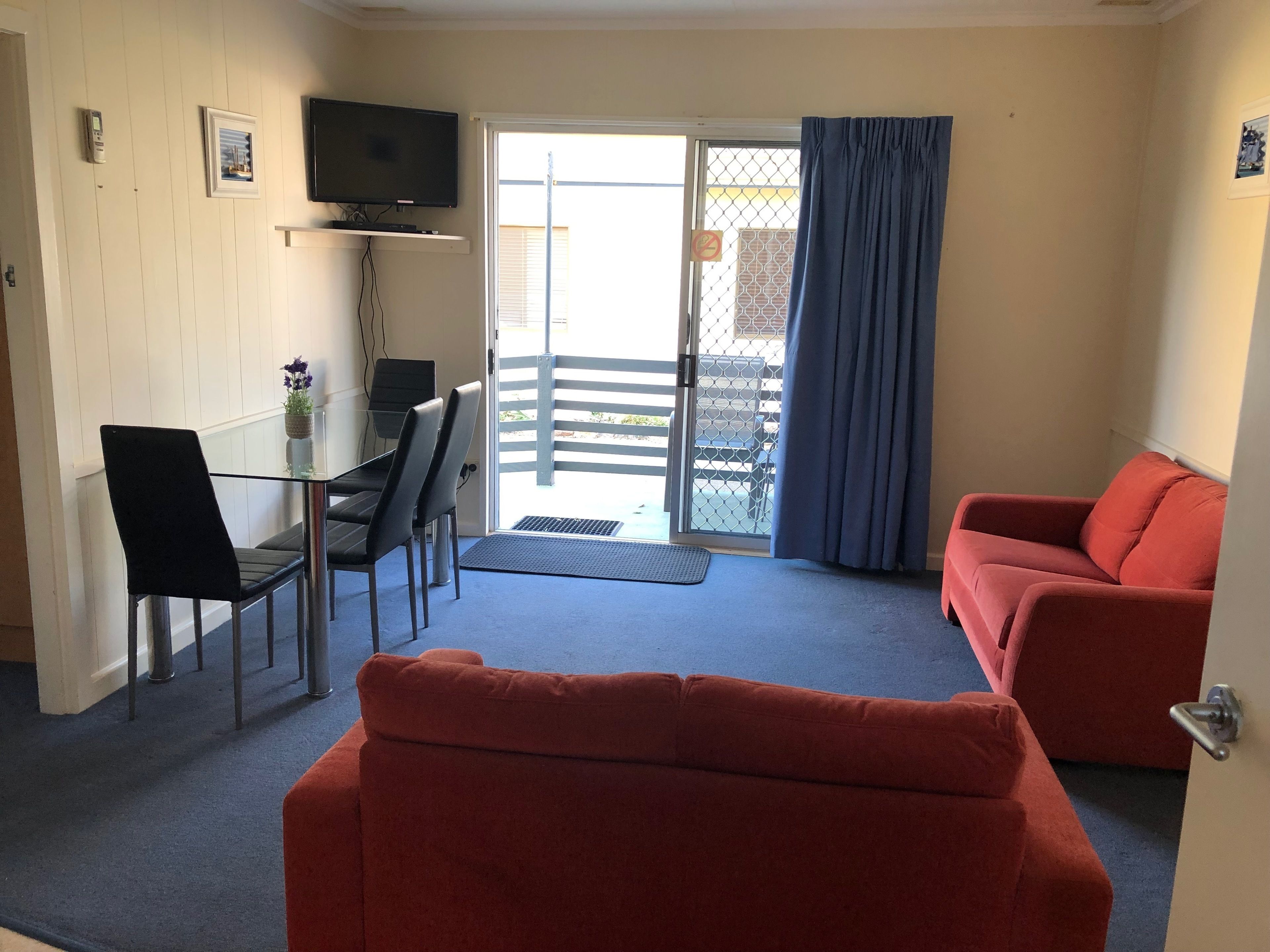 A room with a glass table, black chairs, a red sofa, a TV mounted on the wall, and a sliding glass door leading to an outdoor area.