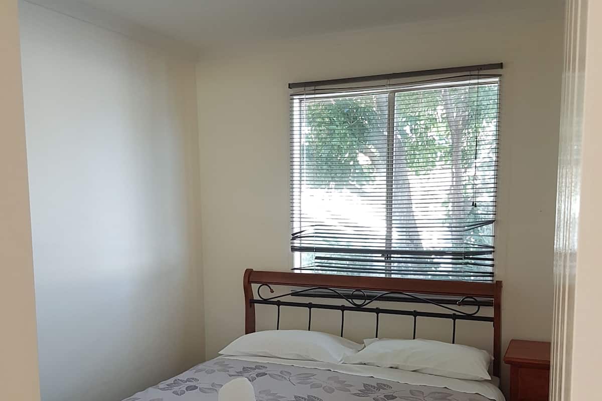 Une petite chambre à coucher, bien aménagée, avec un lit simple, une fenêtre avec des stores, une table de chevet et une porte.