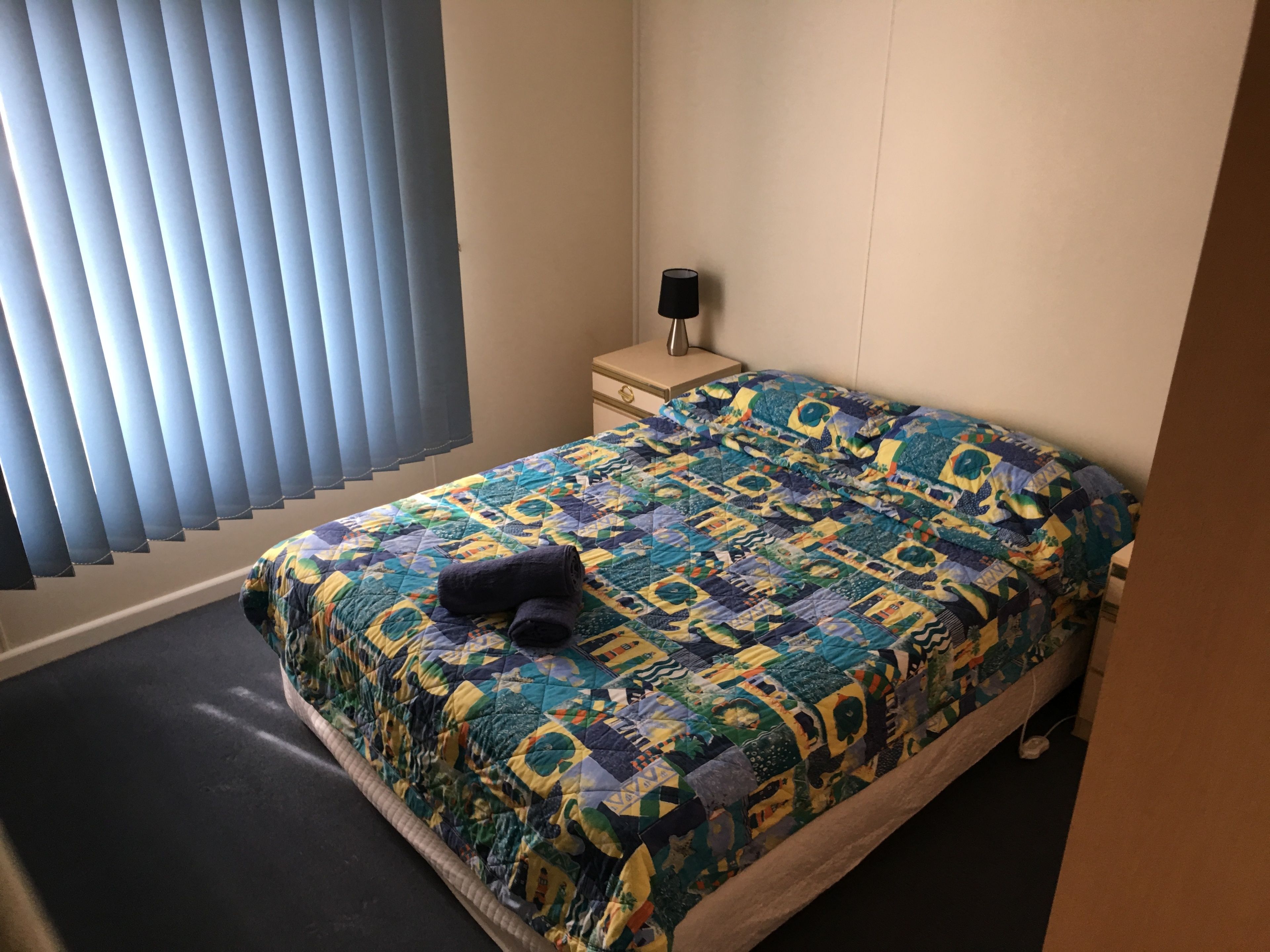 Ein Schlafzimmer mit Bett, Nachttisch, Lampe und einem Fenster mit Jalousien.