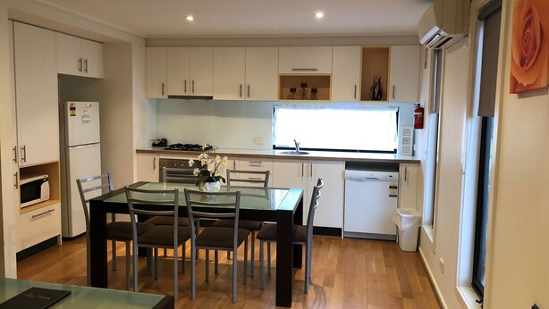 A compact kitchen with white cabinets, a dining table with chairs, a microwave, a refrigerator, and a window with a view. - Koonwarra Holiday Park (Lakes Entrance)