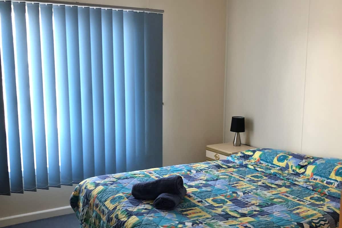 Une chambre d’hôtel avec un grand lit, une table de chevet, une lampe et une fenêtre avec des stores.