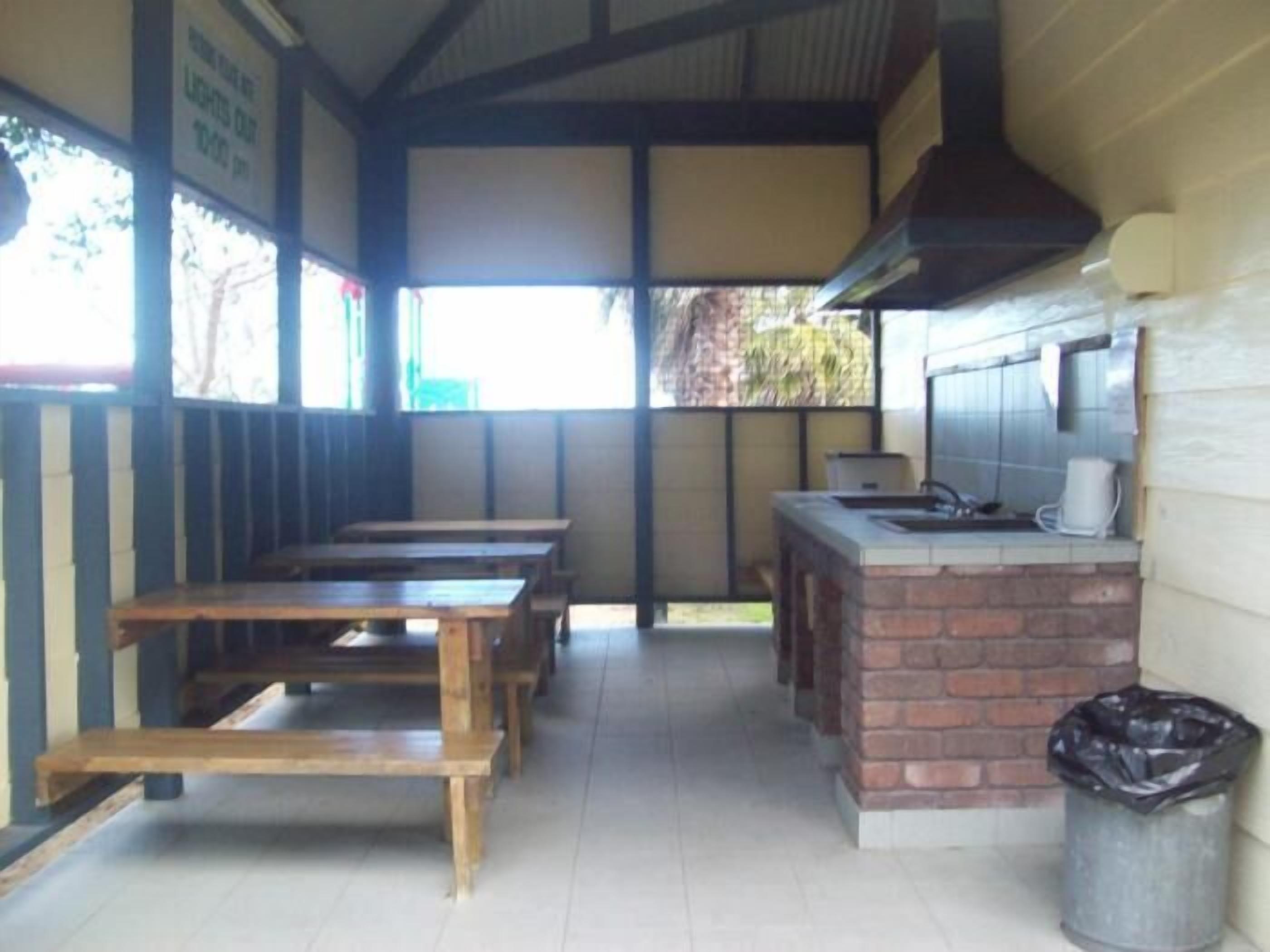 A covered outdoor seating area with wooden tables and benches, a brick counter with a sink, and a trash can.
