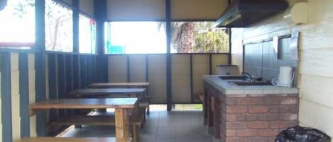A covered outdoor seating area with wooden tables and benches, a brick counter with a sink, and a trash can.
