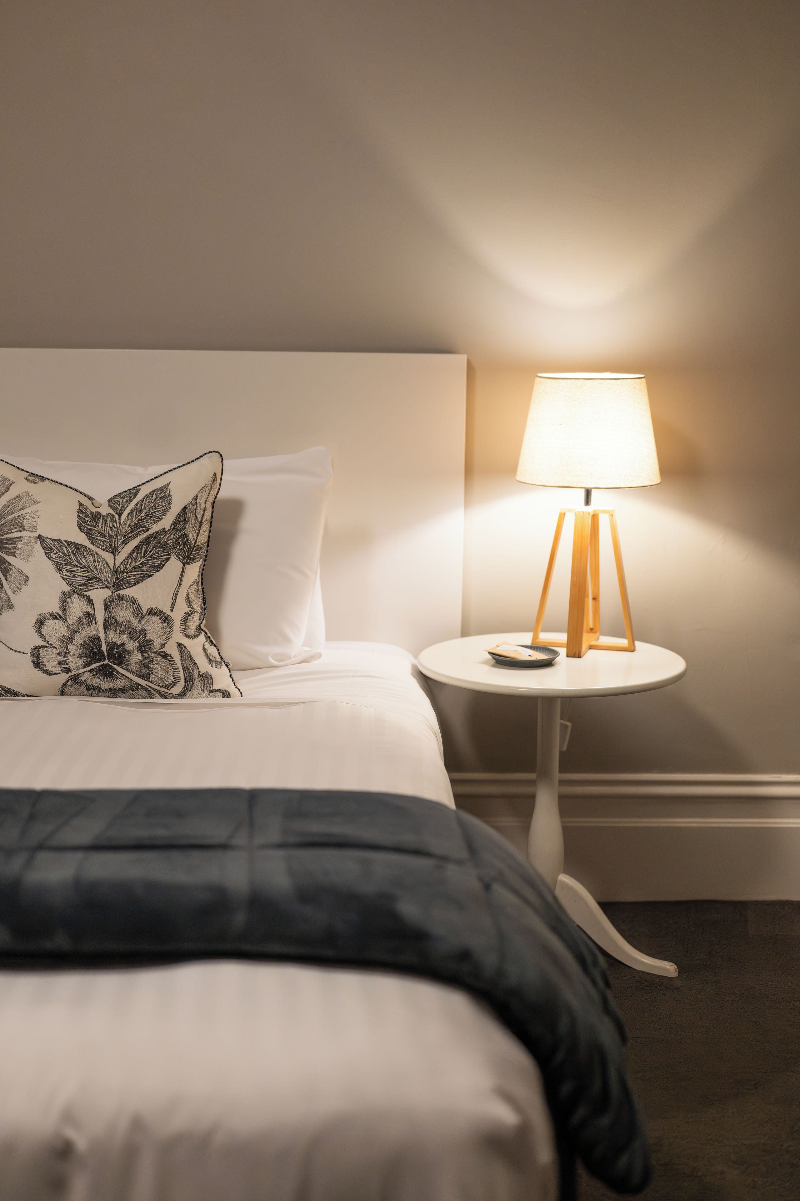 A neatly made bed with a pillow featuring a leaf pattern, a bedside table with a lamp, and a small plate on the table.