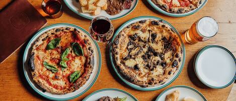 Forskellige retter, herunder pizzaer, salater og grillet kød, anrettet på et træbord.