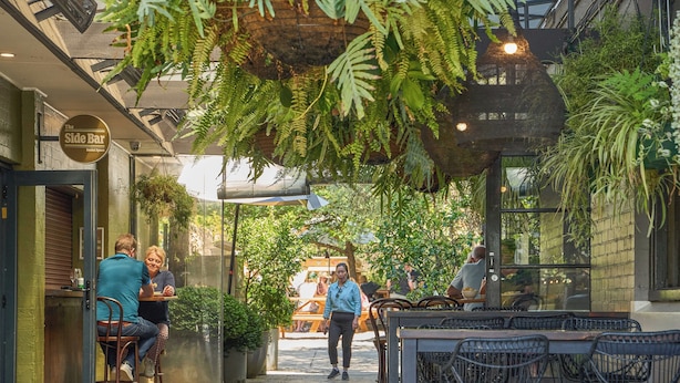 An outdoor dining area with tables and chairs, surrounded by greenery and a brick wall.