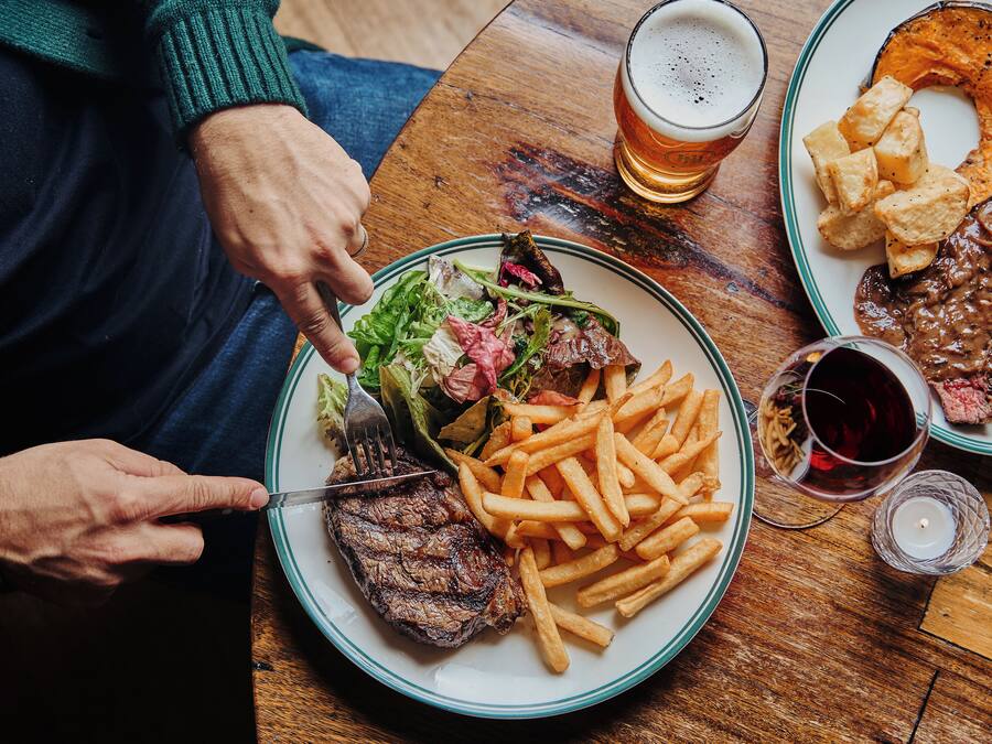 Een eettafel met een bord steak, friet en salade, een glas bier en een kaars.