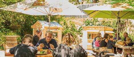 An outdoor dining area with wooden tables and benches, surrounded by trees and greenery.