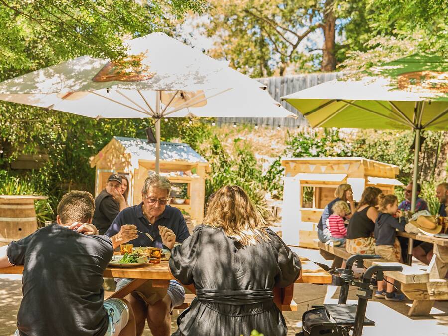 An outdoor dining area with wooden tables and benches, surrounded by trees and greenery.