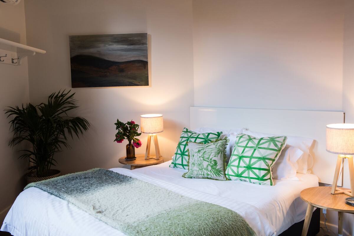 A neatly made bed with a green and white blanket, two bedside lamps, and a painting on the wall.