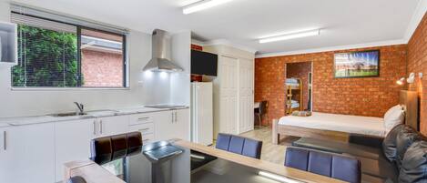 Una cocina moderna con armarios blancos, una mesa de comedor con sillas y un banco empotrado. Cuenta con una pequeña cocina americana con estufa y pileta. La habitación tiene una pared de ladrillo y una ventana con persianas.
