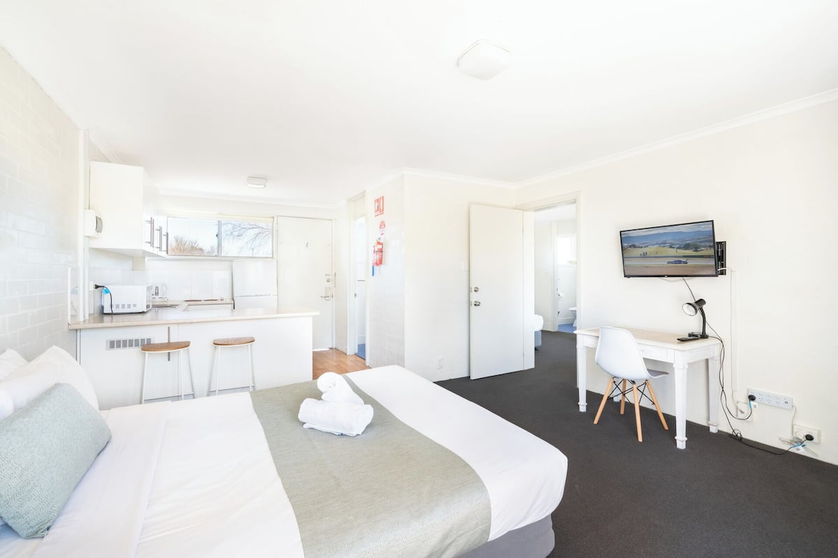 A hotel room with a bed, a desk, a chair, a TV, and a kitchenette.