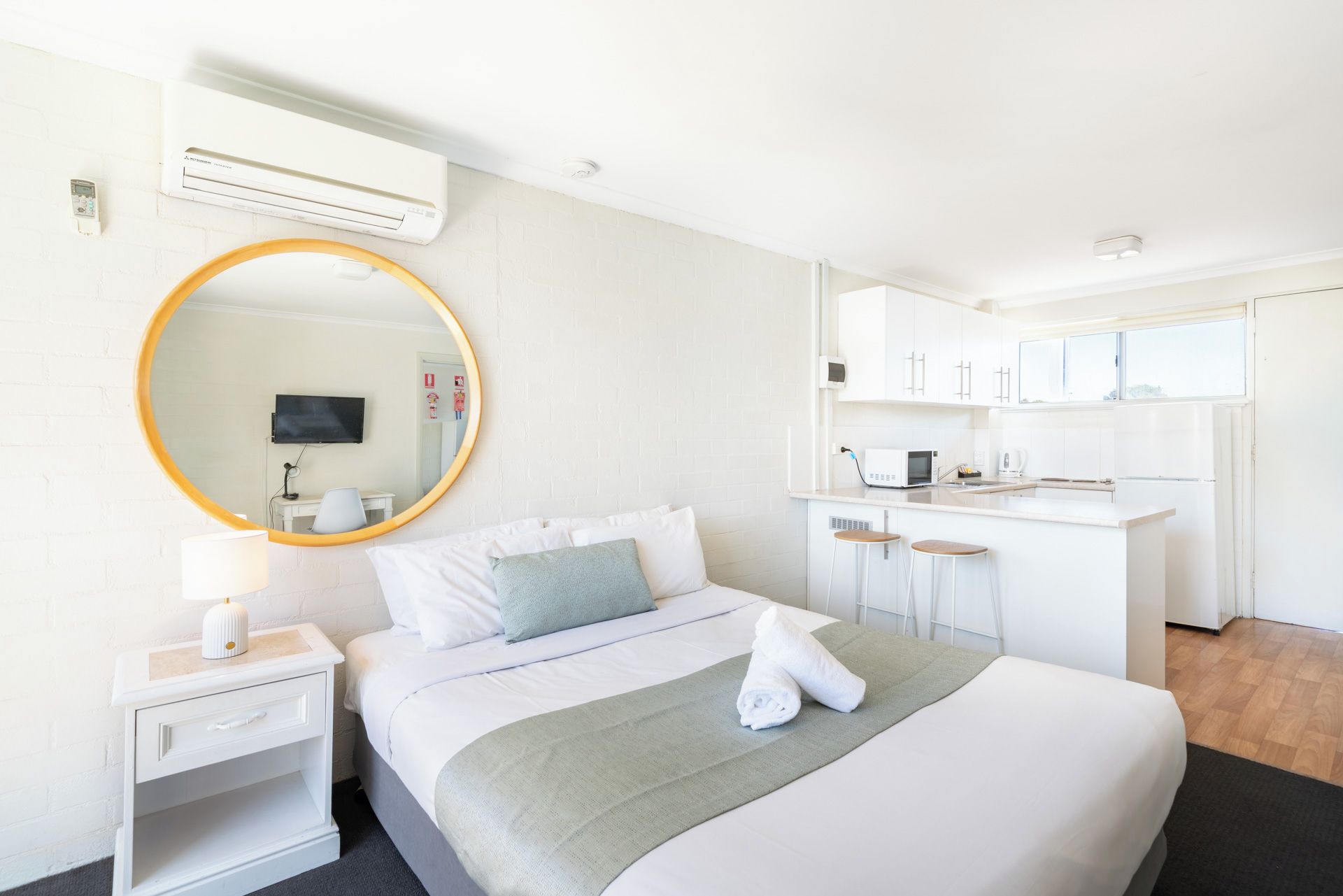 Une chambre d’hôtel comprenant un lit, un miroir rond, une table de chevet, une télévision et une kitchenette équipée d’un four à micro-ondes et d’une machine à café.