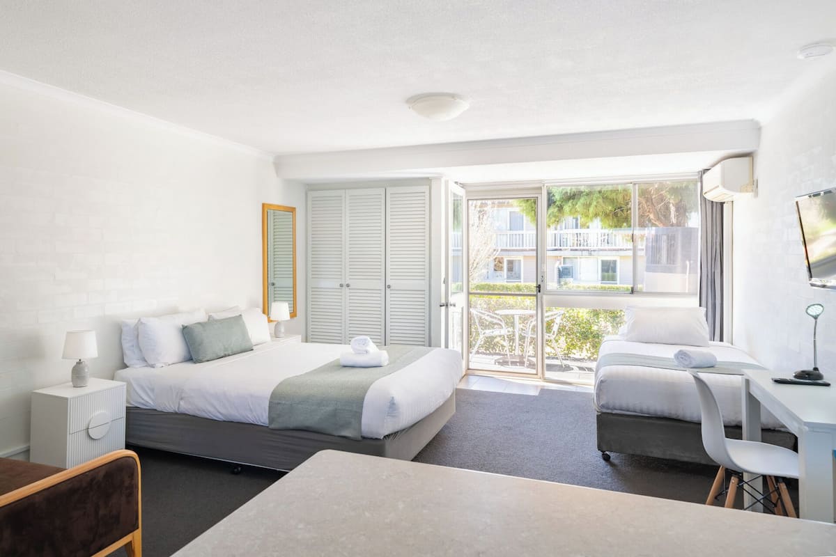 A modern hotel room with a bed, a desk, and a chair. There is a balcony with a view of buildings and trees.