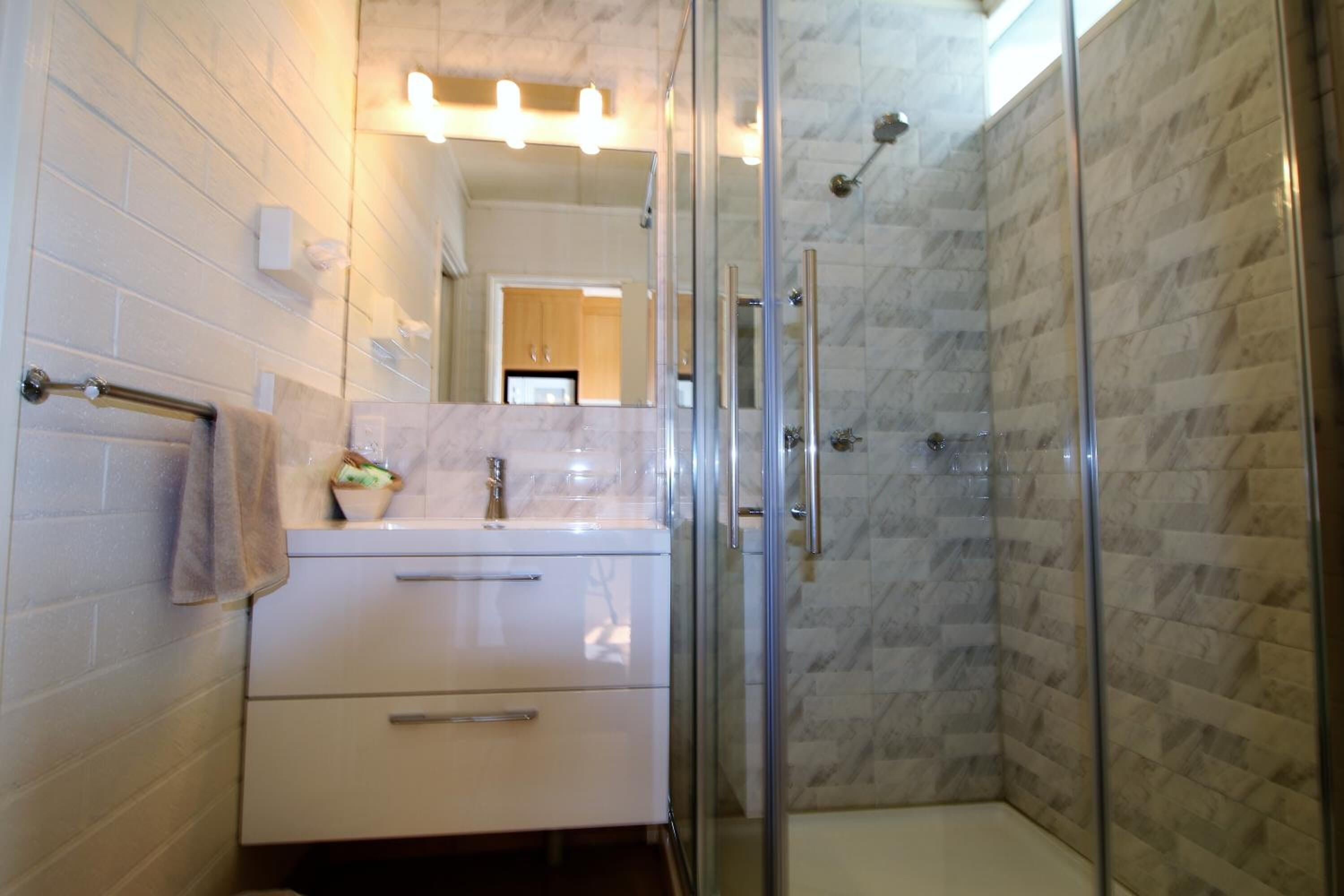 A modern bathroom with a glass shower enclosure, white tiles, and a vanity with a mirror.
