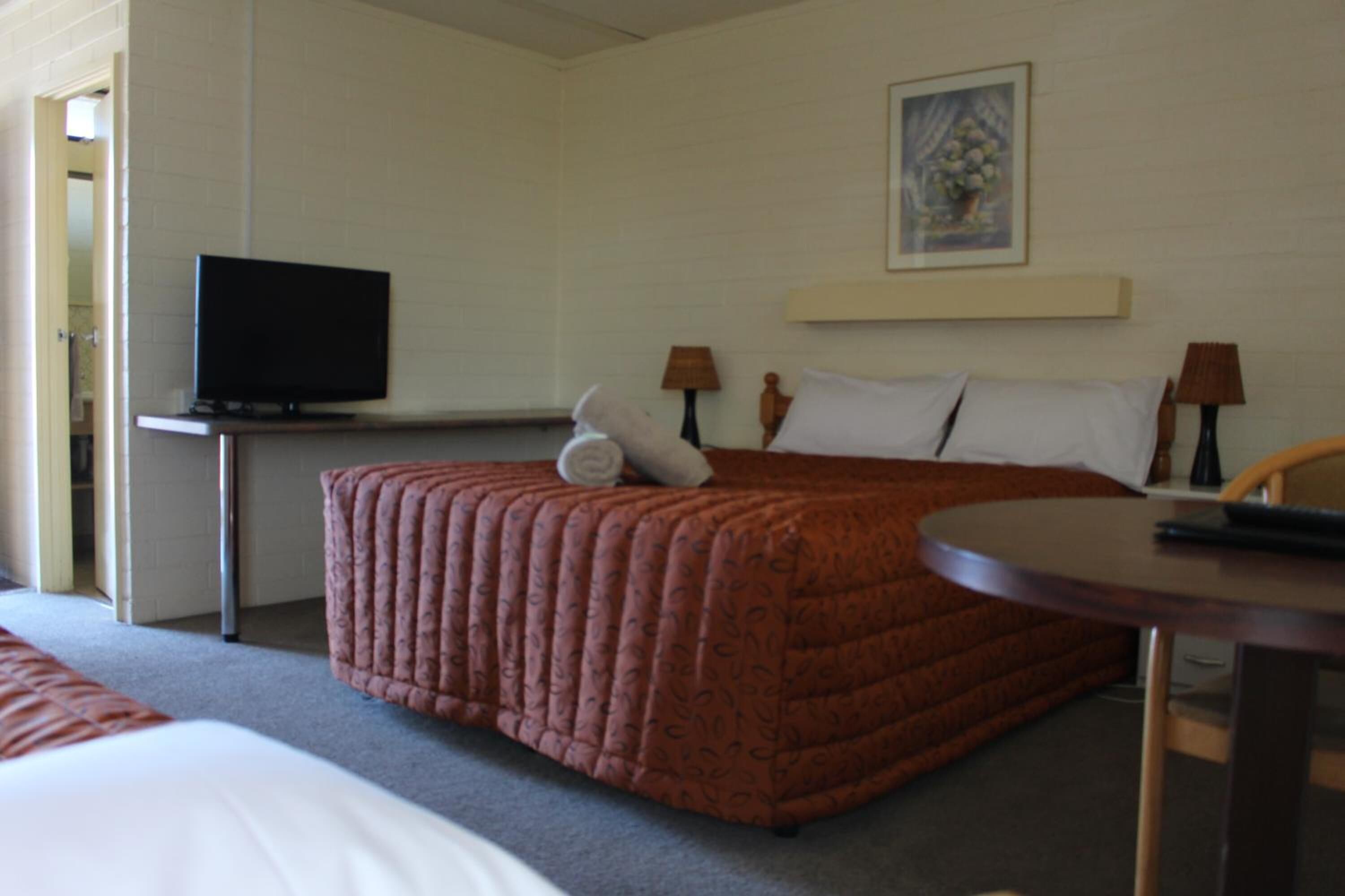 A hotel room with a bed, a television on a stand, a small table, and a painting on the wall.