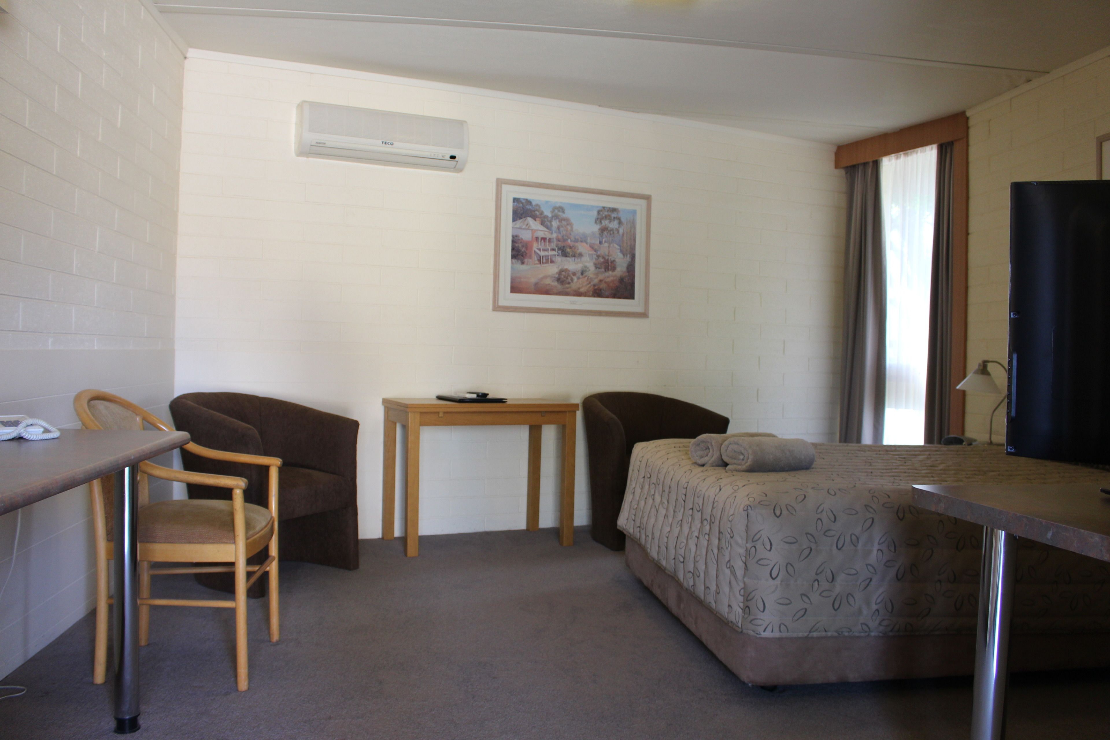 A small, furnished room with a bed, two chairs, a desk, a television, and a painting on the wall.