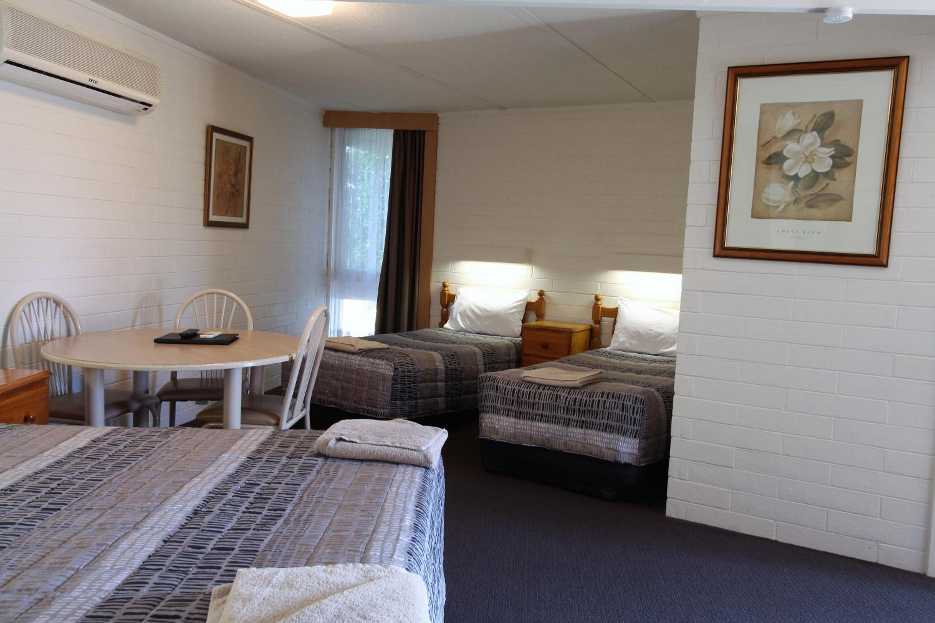 A hotel room with two beds, a round table, and a chair. There is a framed picture on the wall and an air conditioning unit on the wall.