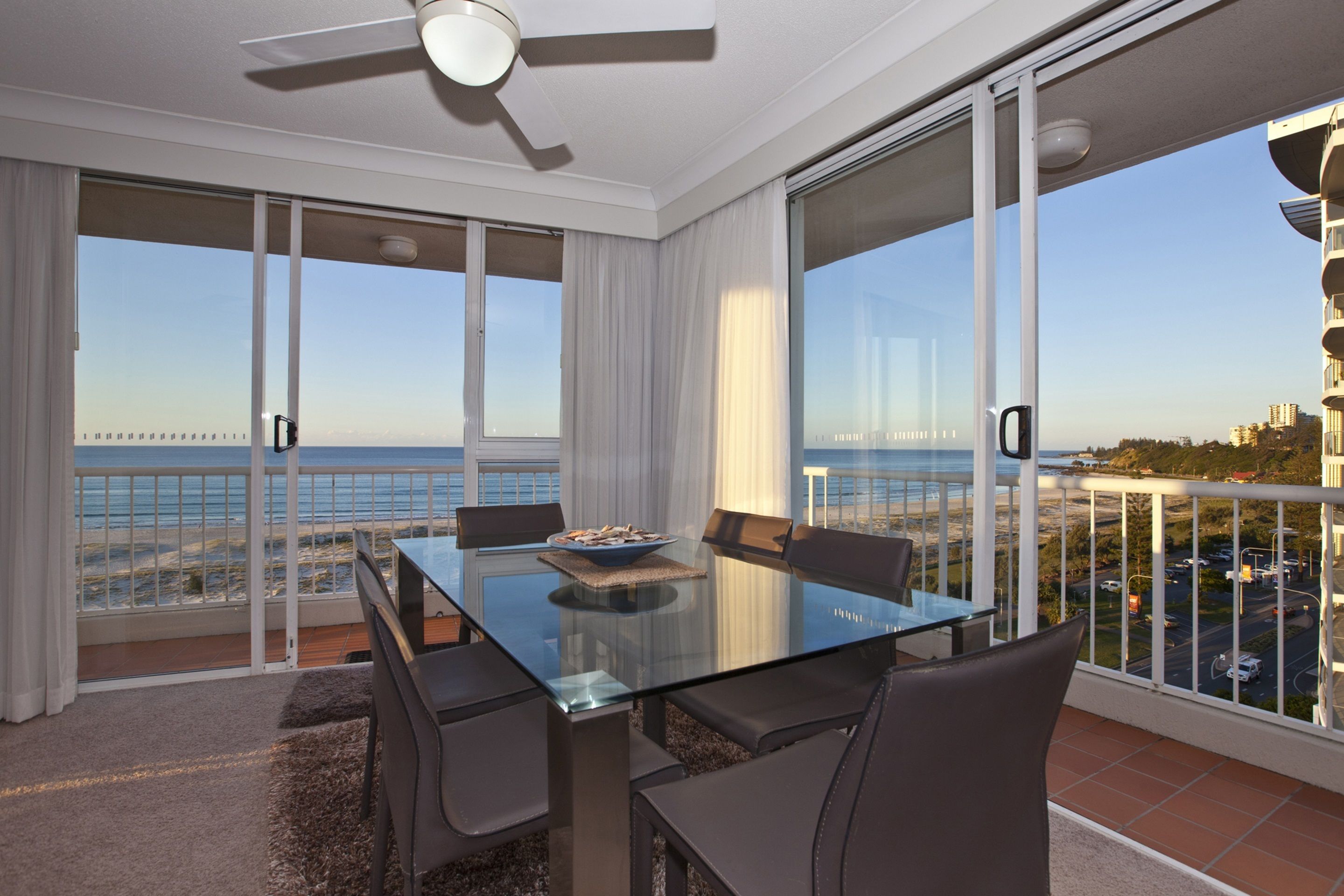 A modern dining area with a glass table, black chairs, and a view of the sea.