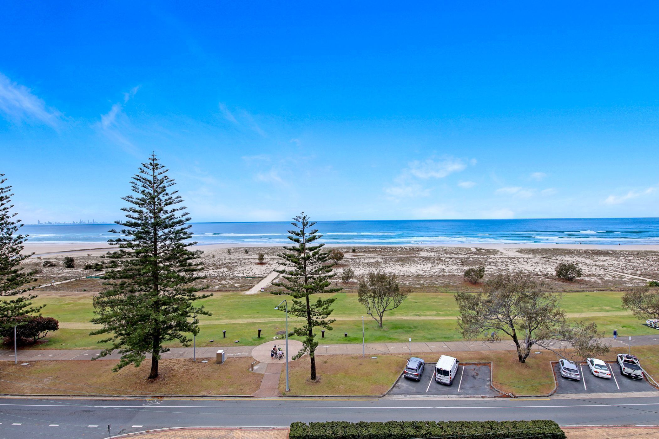 Vista costera con un área de estacionamiento, árboles y una playa al fondo.