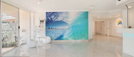 Un pasillo espacioso con un gran mural de una ola, muebles blancos modernos y una pared de vidrio con plantas.