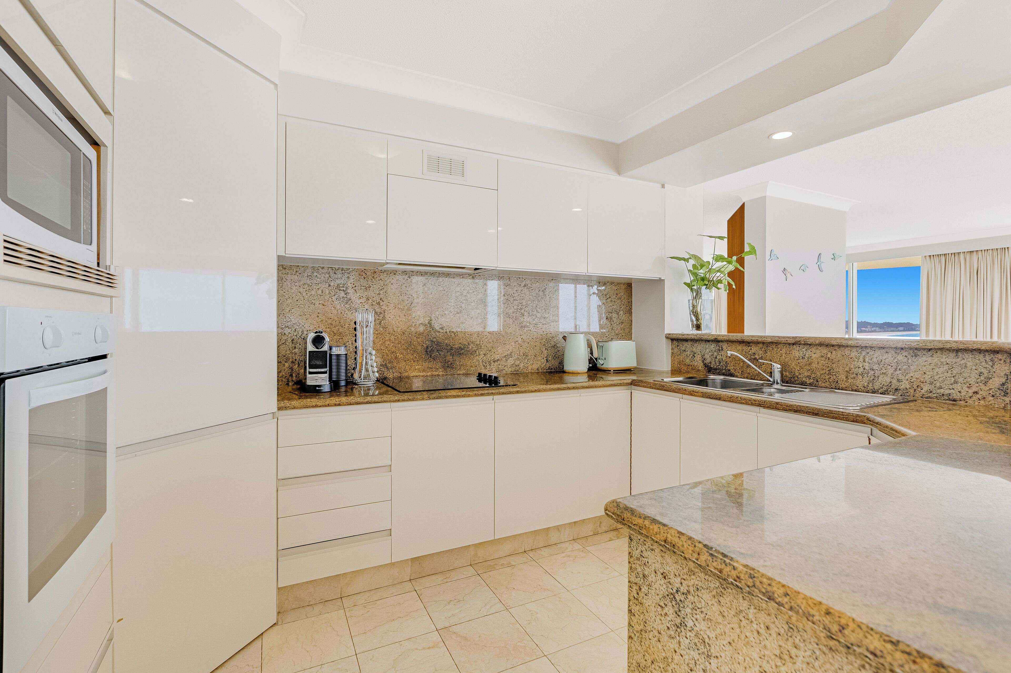 A modern kitchen with white cabinets, granite countertops, a built-in oven, and a large window offering a view.
