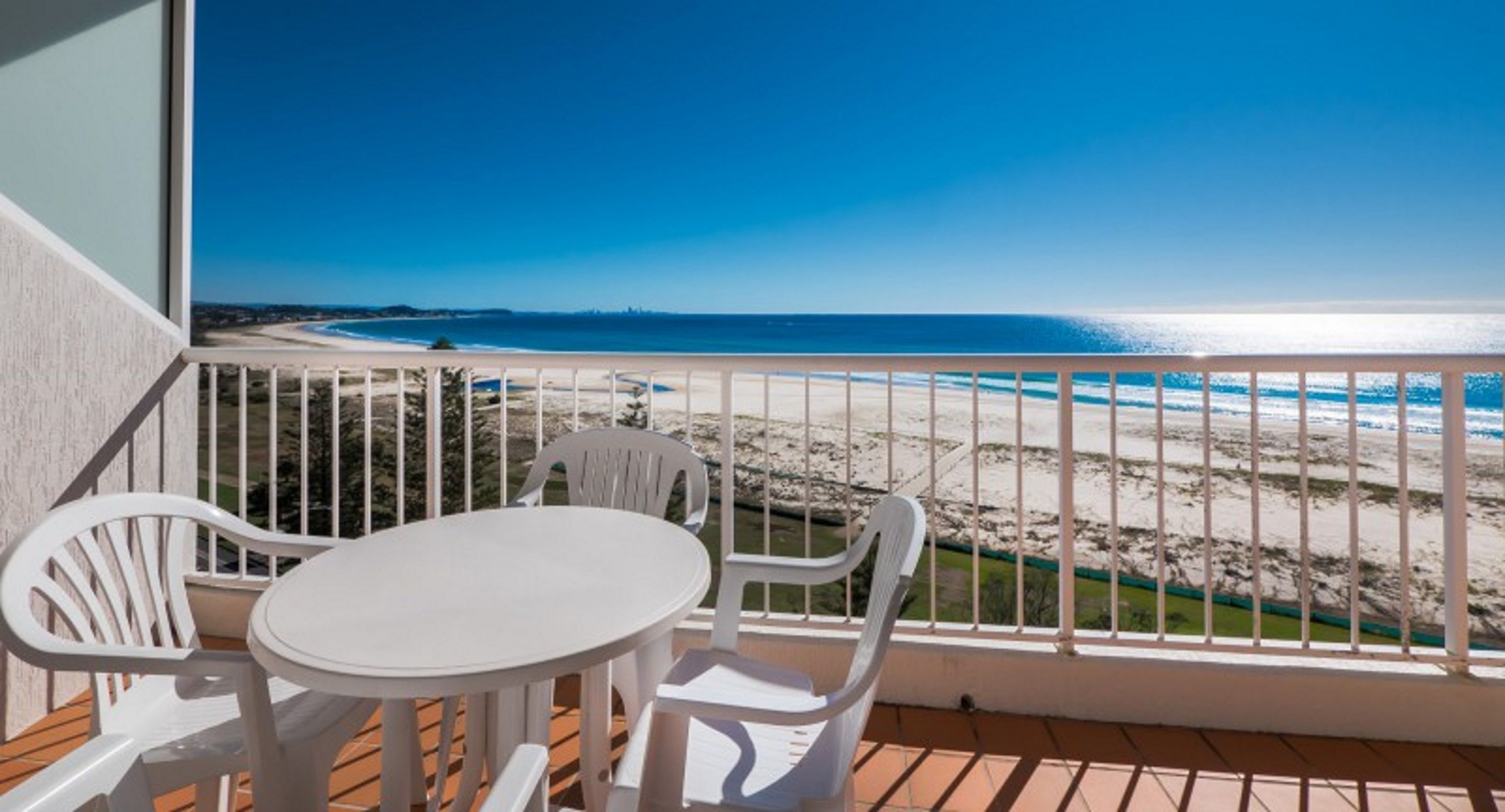 Un balcon avec une table et des chaises blanches, donnant sur une plage sous un ciel bleu azur.