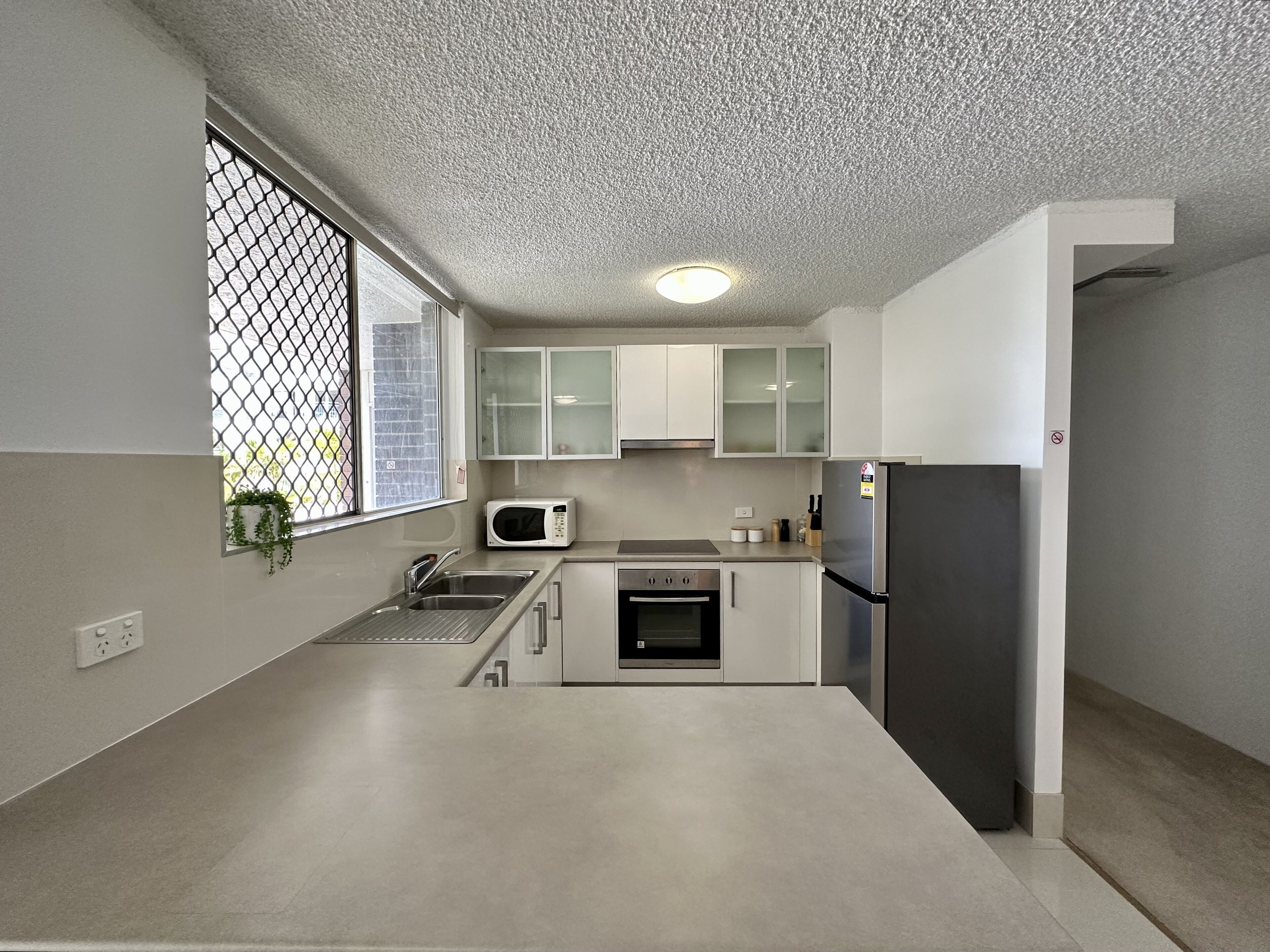 A modern kitchen with a stainless steel refrigerator, a microwave, and a built-in oven. There is a window with a view of the outside, and the kitchen has white cabinets.