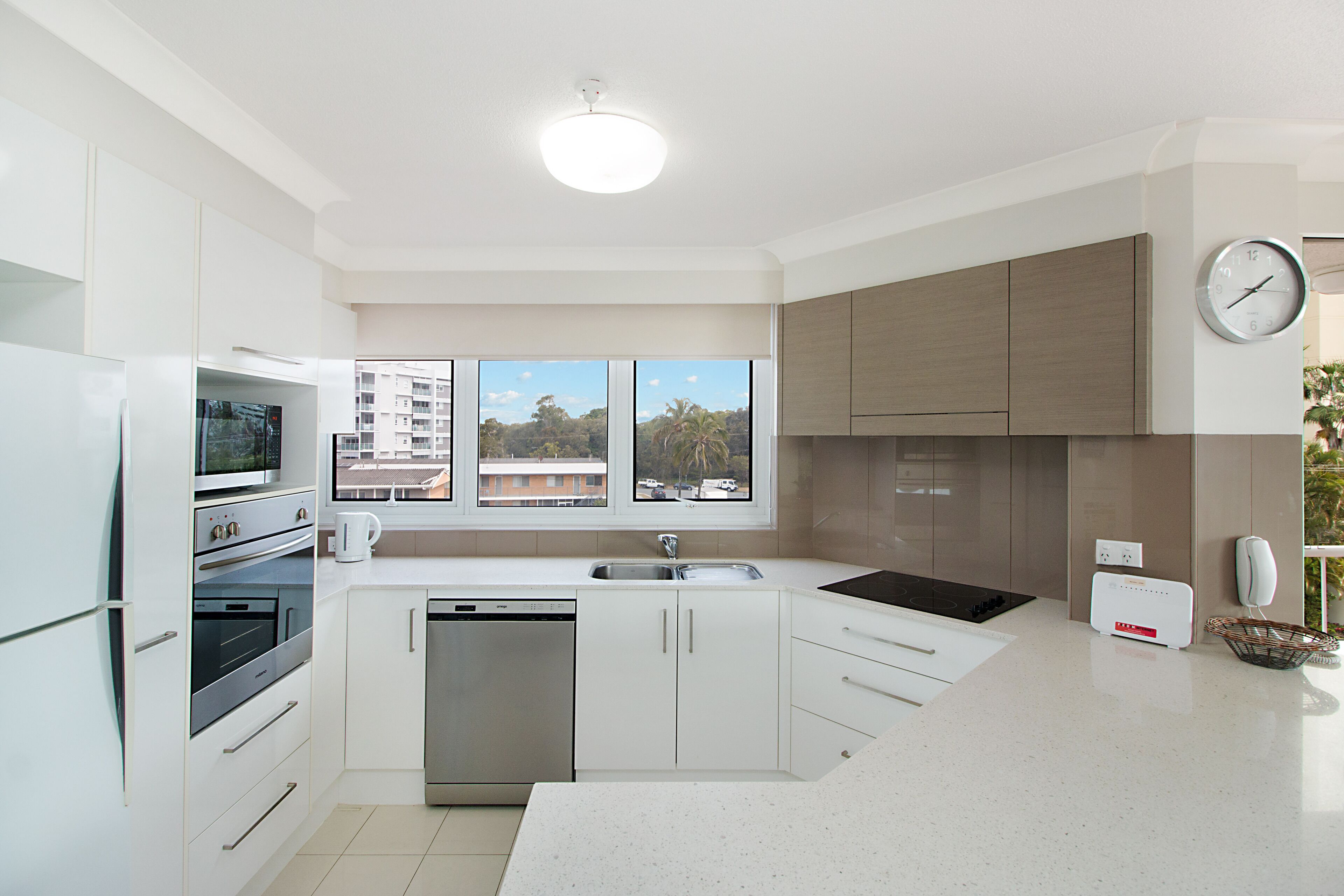 Una cocina moderna con armarios blancos, electrodomésticos de acero inoxidable, un ventanal con vistas a edificios y un reloj en la pared.