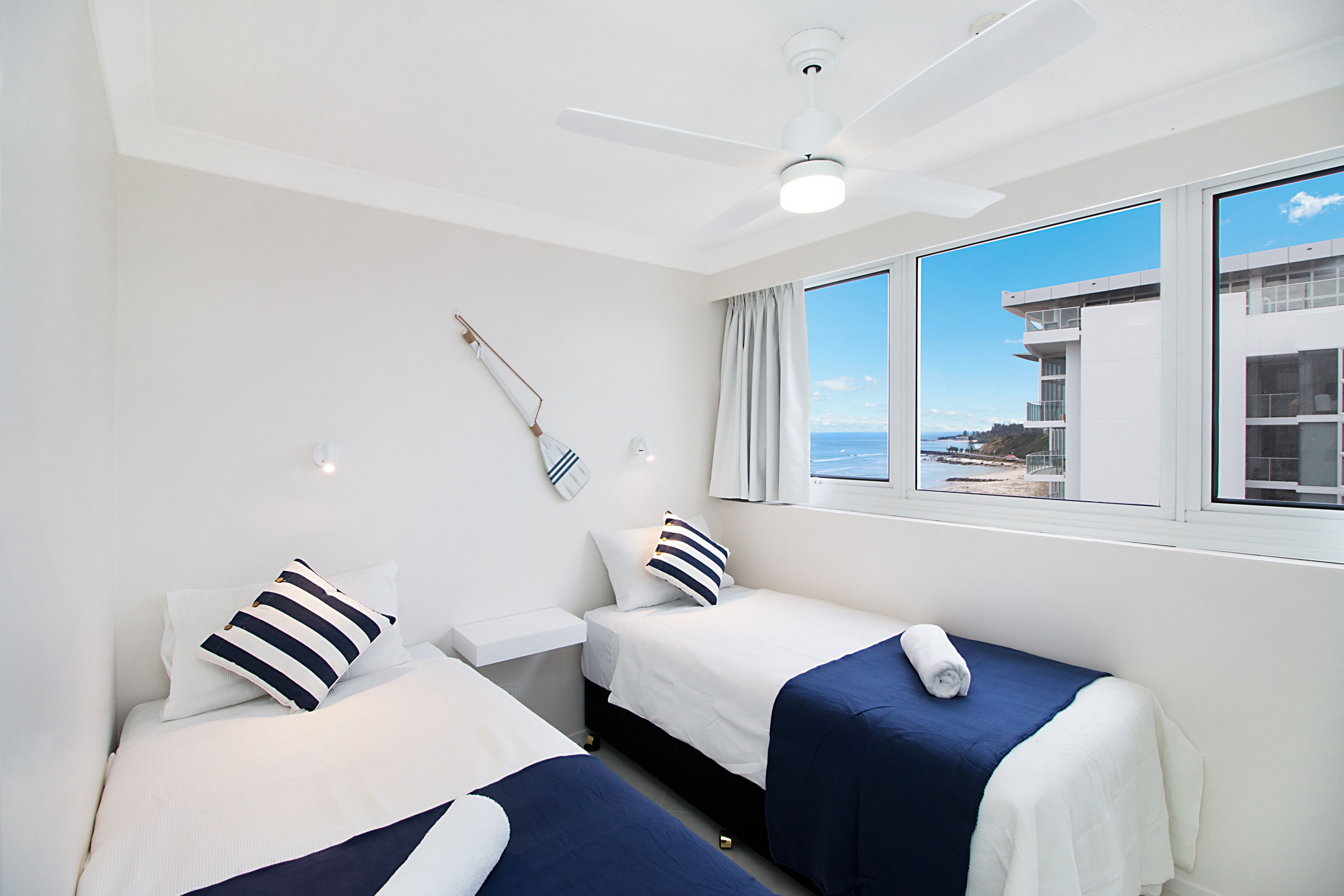 A hotel room with two beds, a ceiling fan, a window with a view of the sea, and a small table with a lamp.