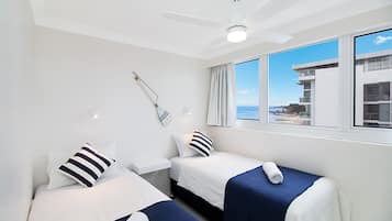 A hotel room with two beds, a ceiling fan, a window with a view of the sea, and a small table with a lamp.