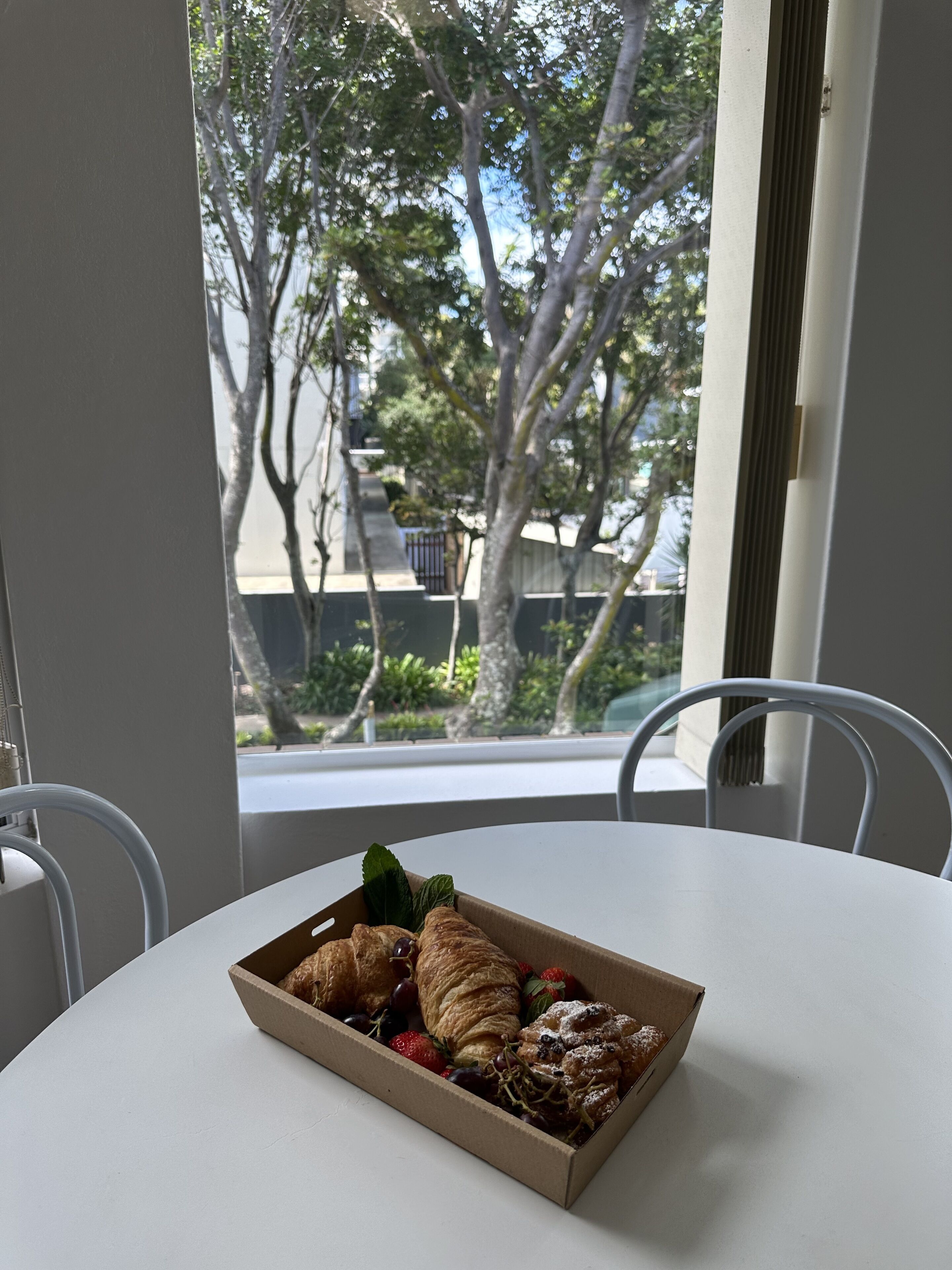 Une table sur laquelle se trouve une boîte en carton contenant des pâtisseries et des fraises, une chaise blanche, et une vue sur des arbres et des bâtiments à travers une fenêtre.