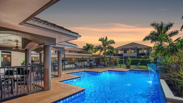 Una piscina con borde de baldosas, rodeada de palmeras y un edificio con balcones.
