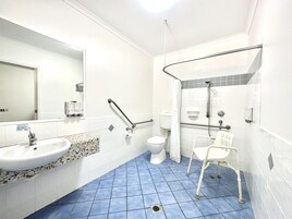 Un baño con lavamanos, inodoro y zona de ducha blanca, que incluye barras de apoyo y una silla accesible para silla de ruedas.